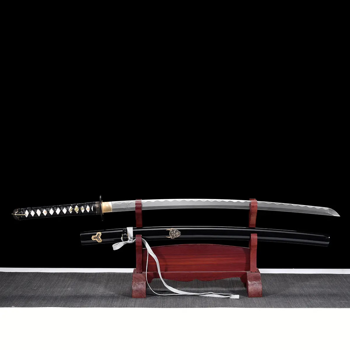 Katana de Bill dans Kill Bill présenté complet sur un support en bois rouge, lame en acier carbone 1045 hamon ondulé, saya noir laqué avec ornements dorés et manche tsuka noir tressé, fond noir minimaliste.