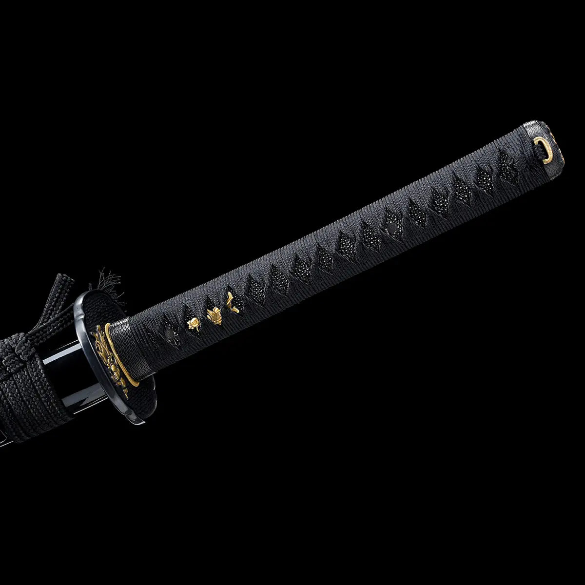 Detailed view of a Japanese katana handle photographed on a deep black background, showing the full length of the black tsuka wrapped in tight black ito over textured ray skin, diamond-shaped grip pattern clearly visible, gold-colored menuki ornaments embedded along the handle, black textured tsuba with gold decorative accents near the guard, emphasizing traditional craftsmanship, balance, and premium samurai sword fittings.
