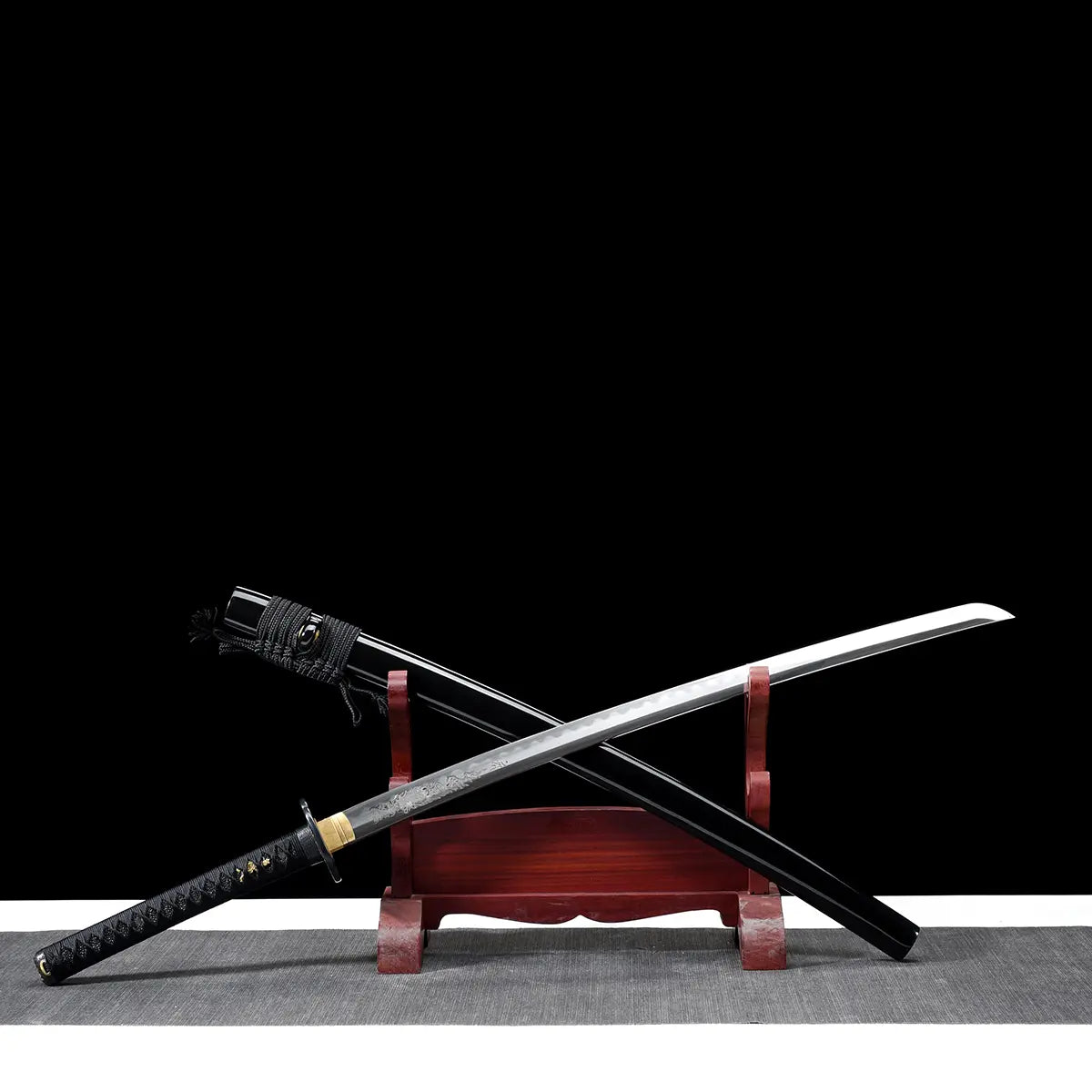 Close-up view of a Japanese katana blade displayed on a dark red wooden stand against a black background, steel blade partially unsheathed showing an engraved figure on the blade near the habaki, black lacquered scabbard positioned behind, golden brass habaki, round black tsuba guard, black handle wrapped with textured ray skin and black cord, fine engraving details clearly visible on the polished steel surface.
