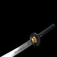 Angled close-up view of a Japanese katana against a deep black background, polished steel blade featuring an engraved Fudo Myoo figure near the base, gold-colored brass habaki fitted tightly against the blade, round black textured tsuba guard visible, black handle wrapped in black cord over ray skin with subtle gold menuki details, focus on blade engraving, fittings, and traditional materials.
