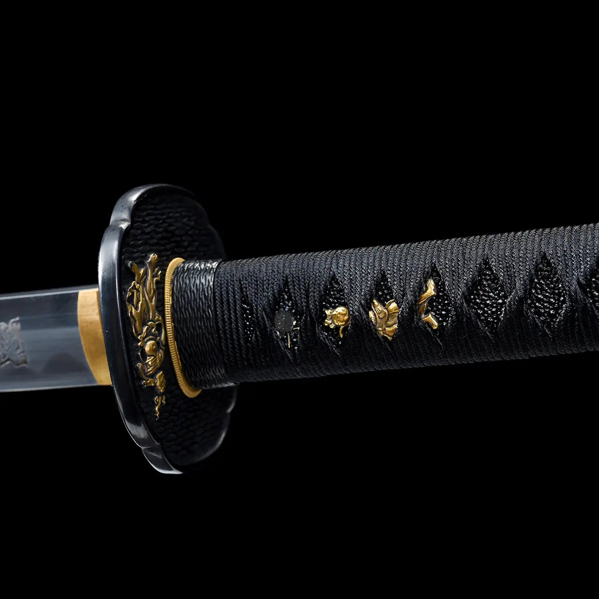Close-up detail of a Japanese katana handle against a deep black background, black tsuka wrapped in tightly woven black cord over textured ray skin, multiple gold-colored menuki figures visible along the grip, round black textured tsuba guard with gold decorative accents, gold brass habaki partially visible at the base of the blade, focus on handle wrapping, ornaments, and traditional fittings.
