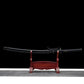 Japanese katana displayed horizontally on a dark red wooden stand against a black background, black lacquered wooden scabbard fitted on the blade, black braided sageo wrapped around the saya, traditional black handle with textured ray skin and black cord wrap, visible round tsuba guard, clean curved blade profile partially concealed by the scabbard.
