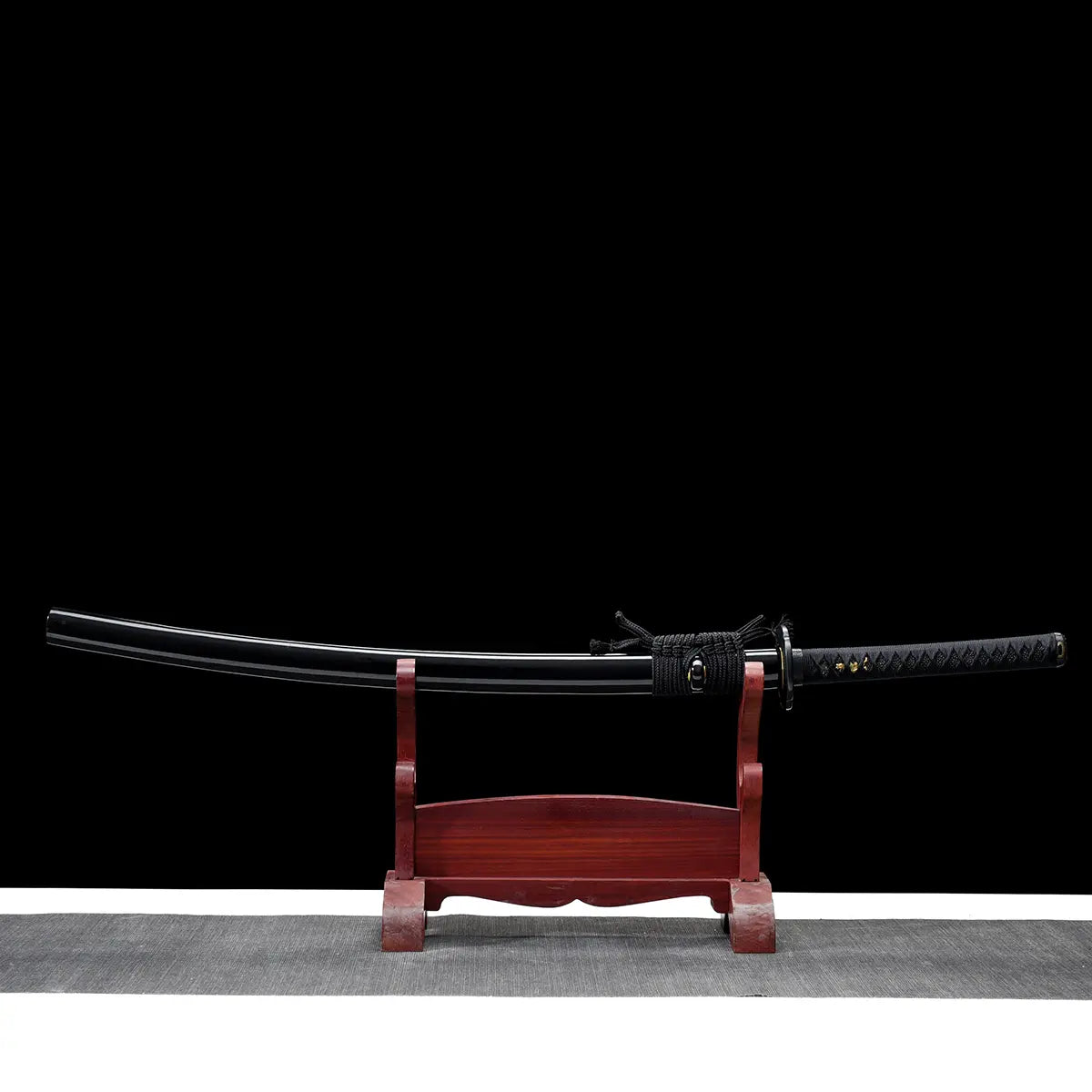 Japanese katana displayed horizontally on a dark red wooden stand against a black background, black lacquered wooden scabbard fitted on the blade, black braided sageo wrapped around the saya, traditional black handle with textured ray skin and black cord wrap, visible round tsuba guard, clean curved blade profile partially concealed by the scabbard.
