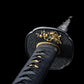 Close-up view of a Japanese katana hilt against a deep black background, black textured tsuba featuring a raised golden relief figure, visible brass habaki ring, black tsuka wrapped in black cord over ray skin, small gold menuki ornaments under the wrap, engraved steel blade fading into the background, sharp focus on traditional fittings and metal textures.
