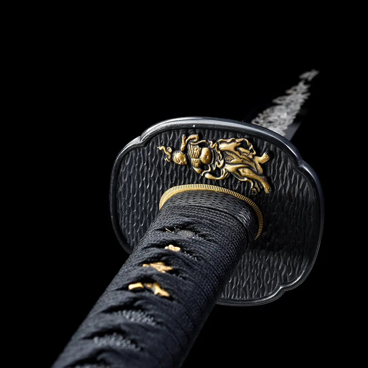 Close-up view of a Japanese katana hilt against a deep black background, black textured tsuba featuring a raised golden relief figure, visible brass habaki ring, black tsuka wrapped in black cord over ray skin, small gold menuki ornaments under the wrap, engraved steel blade fading into the background, sharp focus on traditional fittings and metal textures.
