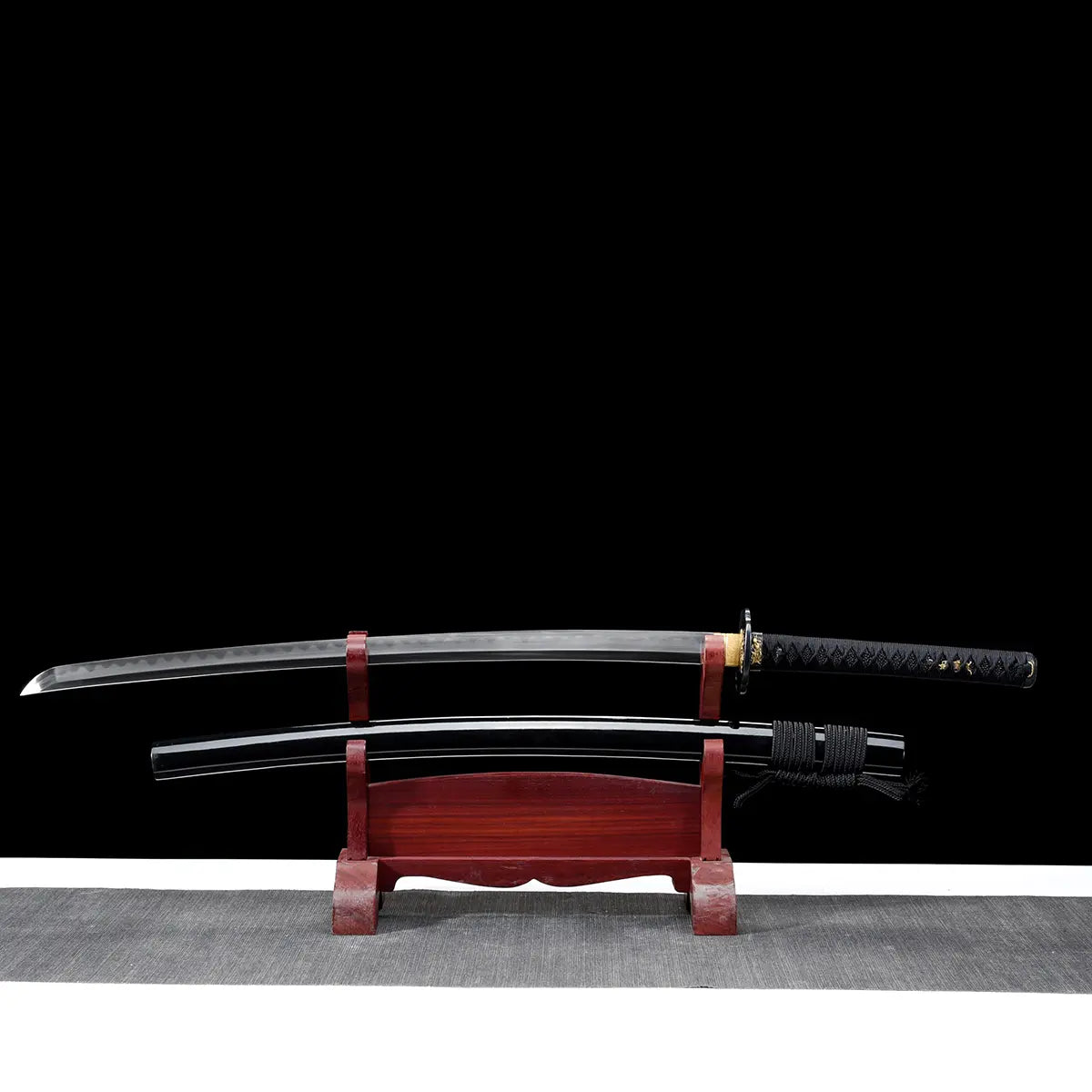 Japanese katana displayed horizontally on a dark red wooden stand against a black background, curved steel blade fully visible without scabbard, polished blade surface with subtle reflections, black handle wrapped in black cord over ray skin, golden habaki near the guard, round black tsuba, traditional katana silhouette resting above the black lacquered scabbard placed below on the stand.
