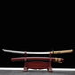 Katana Himura Kenshin Sakabato a lame inversee expose sur un support en bois rouge devant un fond noir, sabre degainne avec lame courbee en acier, fourreau rouge laque pose sous le katana, manche beige tresse avec peau de raie et garde noire ronde.
