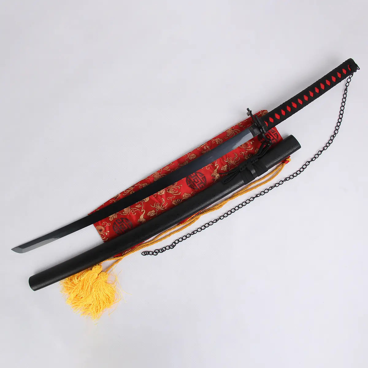 Katana Ichigo Kurosaki Tensa Zangetsu de Bleach, lame partiellement sortie de son fourreau noir mat avec chaîne, manche noir et rouge, présenté sur un sac en soie rouge à motifs dorés avec pompon jaune, fond blanc clair.