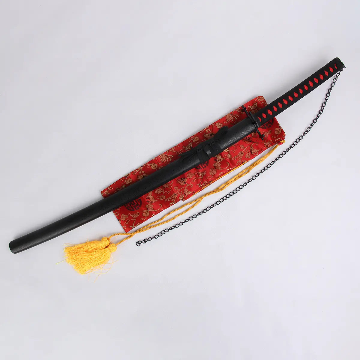 Photo du katana Ichigo Kurosaki Tensa Zangetsu de Bleach, lame dans son fourreau noir mat avec chaîne, posé sur un sac de soie rouge à motifs dorés avec pompon jaune, fond blanc neutre.