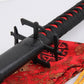 Détail du katana Ichigo Kurosaki Tensa Zangetsu de Bleach, garde noire en croix (tsuba) et tsuka noir tressé avec fond rouge, reposant sur un sac en soie rouge à motifs dorés, fond blanc lumineux.