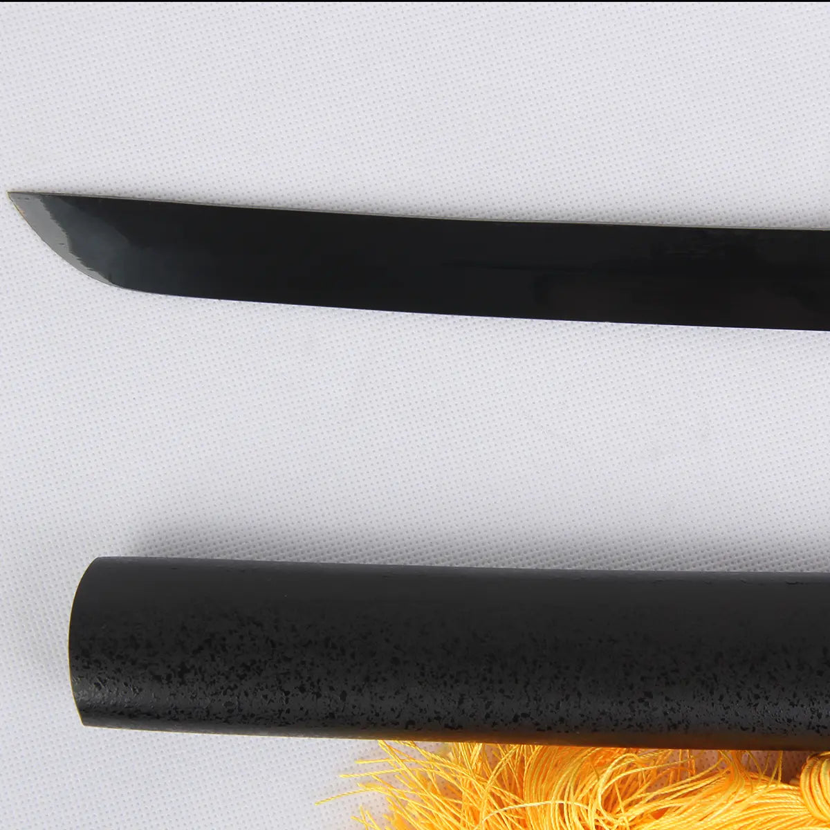 Gros plan de la lame noire du katana Ichigo Kurosaki Tensa Zangetsu de Bleach, partiellement sortie de son fourreau noir texturé, posé sur un sac en soie rouge avec pompon jaune, fond blanc neutre.