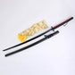 Réplique du katana Ichigo Kurosaki Tensa Zangetsu version classique, inspiré du Bankai de Bleach, lame en acier carbone 1045, avec fourreau noir mat, poignée noire tressée à losanges rouges, chaînette décorative et housse dorée posés sur fond blanc.