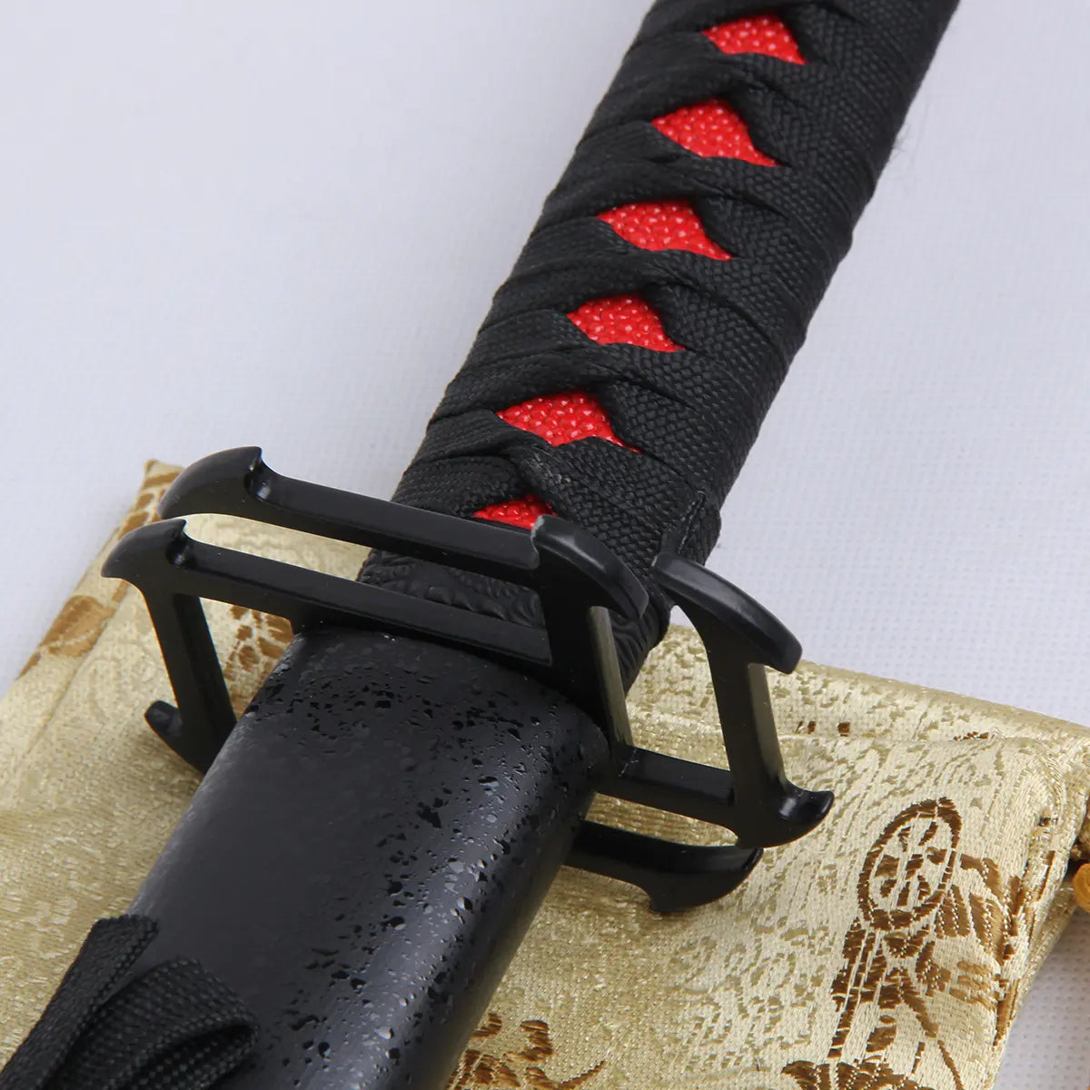 Garde carrée noire du katana Ichigo Kurosaki Tensa Zangetsu version stylisée, réplique collector Bleach, avec poignée noire tressée révélant un fond rouge, posée sur un tissu doré orné de motifs japonais.
