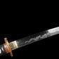 Détail de la lame du katana japonais artisanal Ryū no Kiba sur fond noir uni, lame sortie du fourreau montrant un horimono représentant un dragon enroulé autour d’un trident, hamon ondulé visible sur l’acier poli, habaki doré ajusté contre la garde tsuba, poignée tsuka noire tressée partiellement visible sans support.
