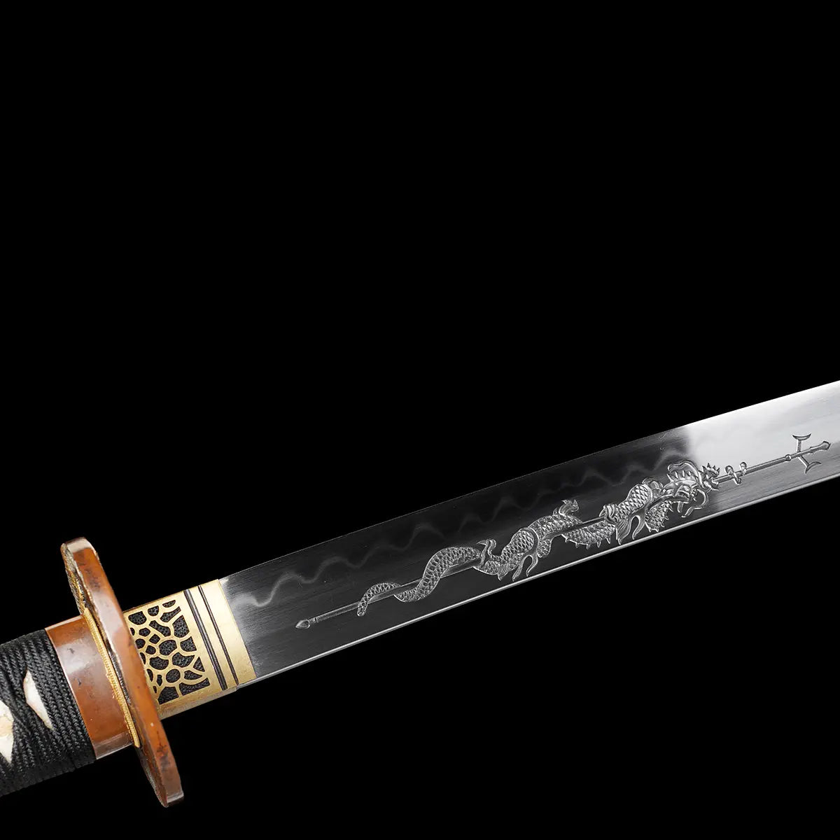 Détail de la lame du katana japonais artisanal Ryū no Kiba sur fond noir uni, lame sortie du fourreau montrant un horimono représentant un dragon enroulé autour d’un trident, hamon ondulé visible sur l’acier poli, habaki doré ajusté contre la garde tsuba, poignée tsuka noire tressée partiellement visible sans support.

