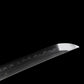 Gros plan sur la pointe de la lame du katana japonais artisanal Ryū no Kiba sur fond noir uni, acier poli montrant un hamon ondulé visible le long du tranchant, pointe kissaki nette et affûtée, surface de la lame lisse sans fourreau ni support apparent.
