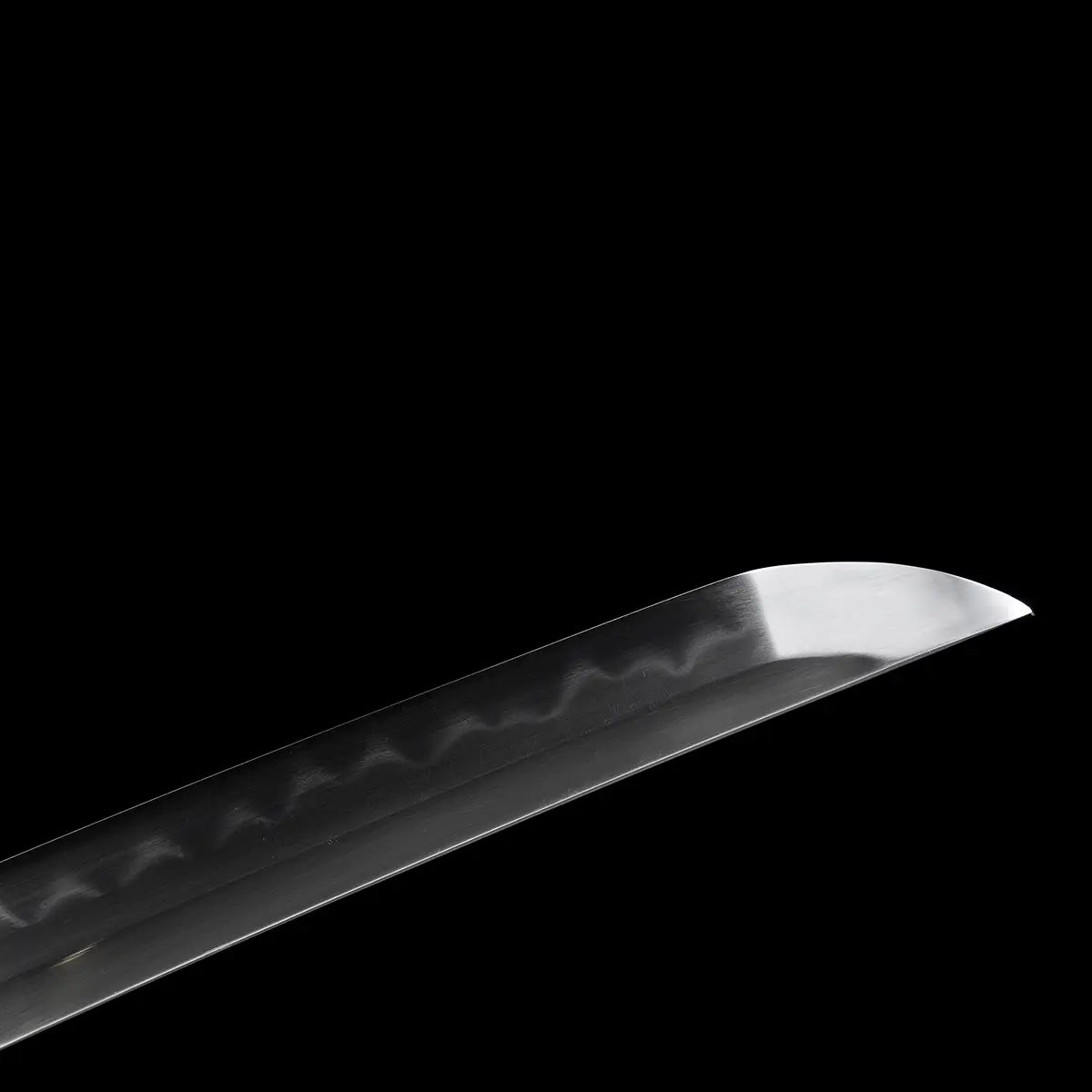 Gros plan sur la pointe de la lame du katana japonais artisanal Ryū no Kiba sur fond noir uni, acier poli montrant un hamon ondulé visible le long du tranchant, pointe kissaki nette et affûtée, surface de la lame lisse sans fourreau ni support apparent.
