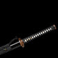 Détail de la poignée du katana japonais artisanal Ryū no Kiba sur fond noir uni, tsuka longue enveloppée d’un tressage noir avec losanges blancs en peau de raie et menuki dorés visibles, garde tsuba ronde en métal patiné br
