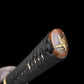 Gros plan sur l’extrémité de la poignée du katana japonais artisanal Ryū no Kiba sur fond noir uni, kashira en métal patiné brun ornée d’un motif décoratif doré, tsuka enveloppée d’un tressage noir serré avec losanges blancs en peau de raie, texture du cordage bien visible, aucune lame, fourreau ou support apparent.

