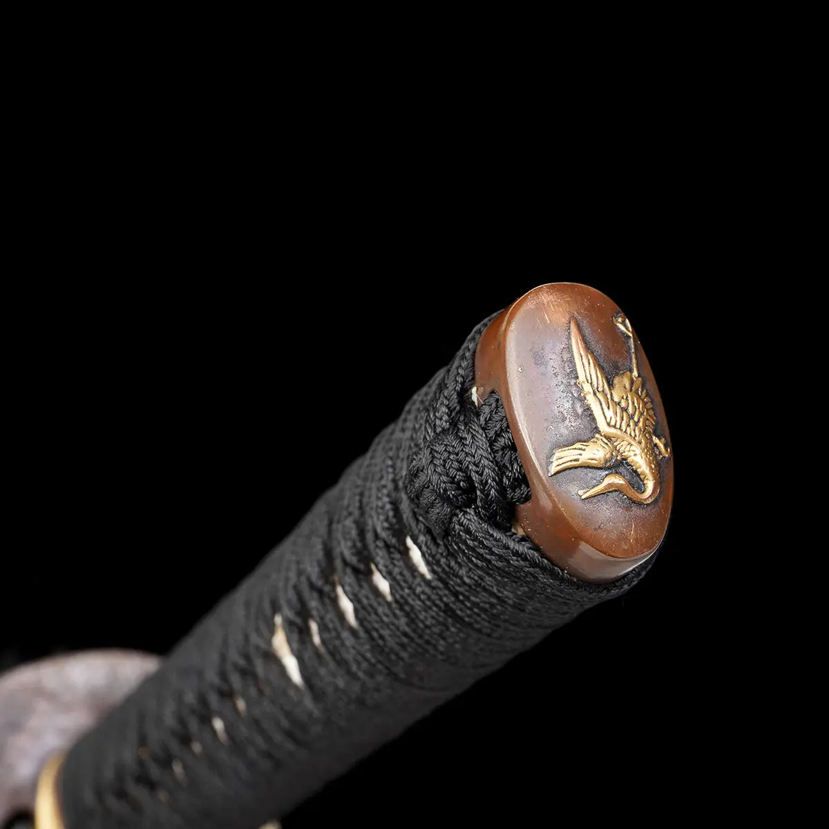 Gros plan sur l’extrémité de la poignée du katana japonais artisanal Ryū no Kiba sur fond noir uni, kashira en métal patiné brun ornée d’un motif décoratif doré, tsuka enveloppée d’un tressage noir serré avec losanges blancs en peau de raie, texture du cordage bien visible, aucune lame, fourreau ou support apparent.
