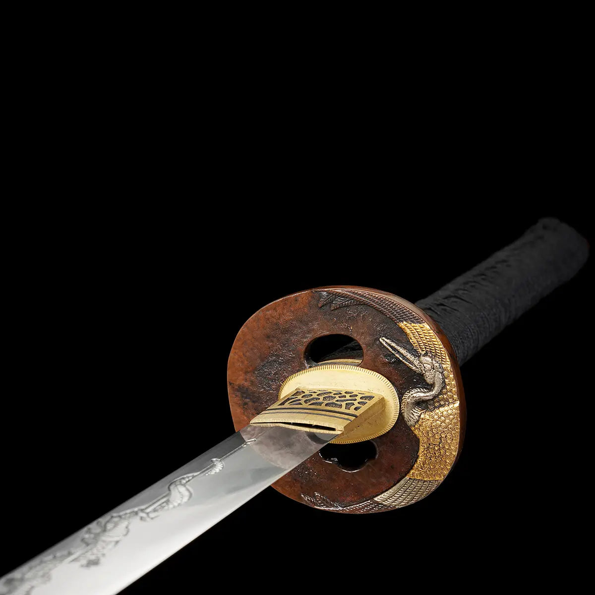 Gros plan sur la garde du katana japonais artisanal Ryū no Kiba sur fond noir uni, tsuba ronde en métal patiné brun avec motifs décoratifs et éléments dorés représentant un dragon, habaki doré finement ajusté, départ de la lame en acier poli visible, poignée tsuka noire tressée partiellement visible, aucun support apparent.
