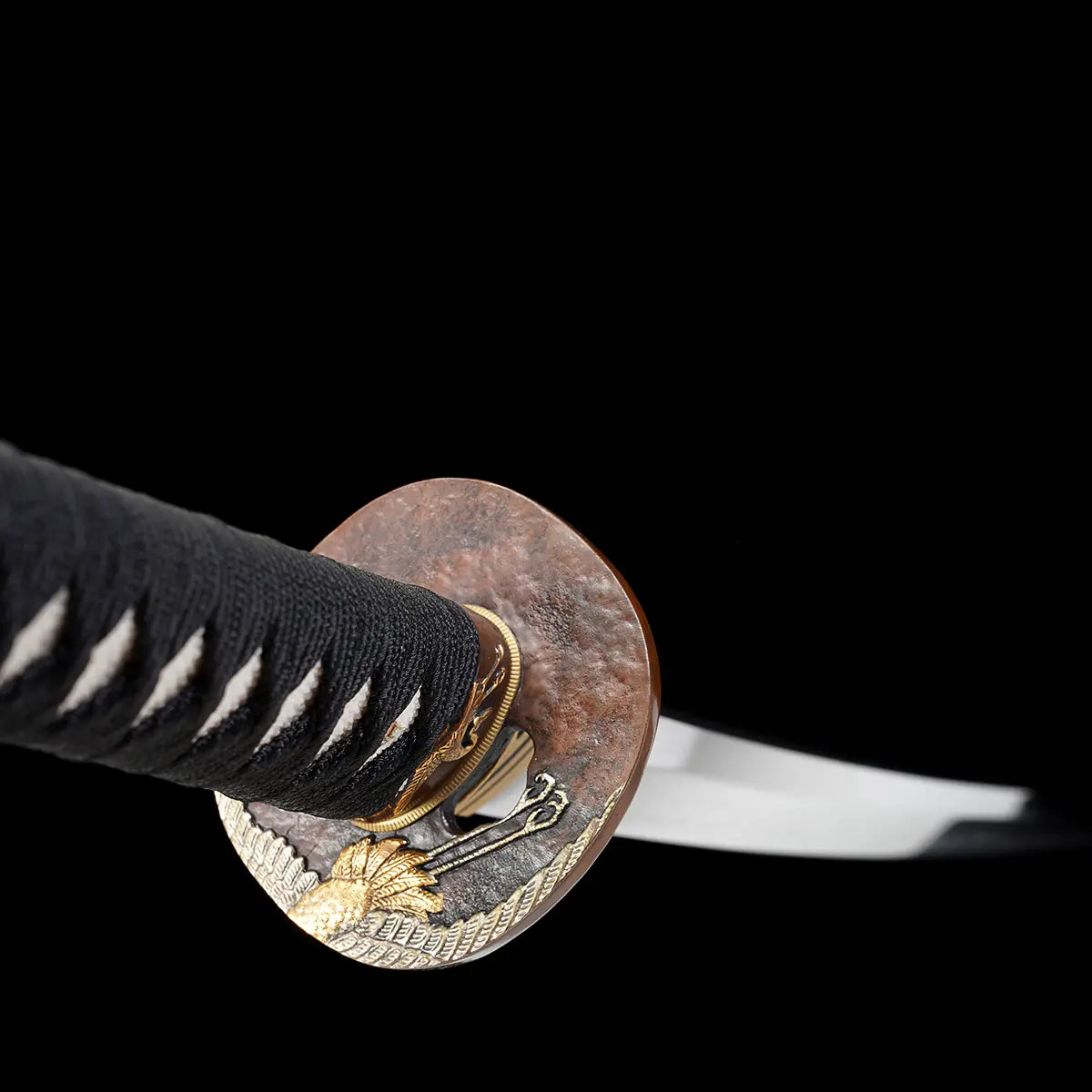 Gros plan sur la garde du katana japonais artisanal Ryū no Kiba sur fond noir uni, tsuba ronde en métal patiné brun avec motifs décoratifs dorés, habaki doré ajusté à la base de la lame, poignée tsuka noire tressée avec losanges blancs en peau de raie visibles, lame légèrement floutée en arrière-plan, aucun support apparent.
