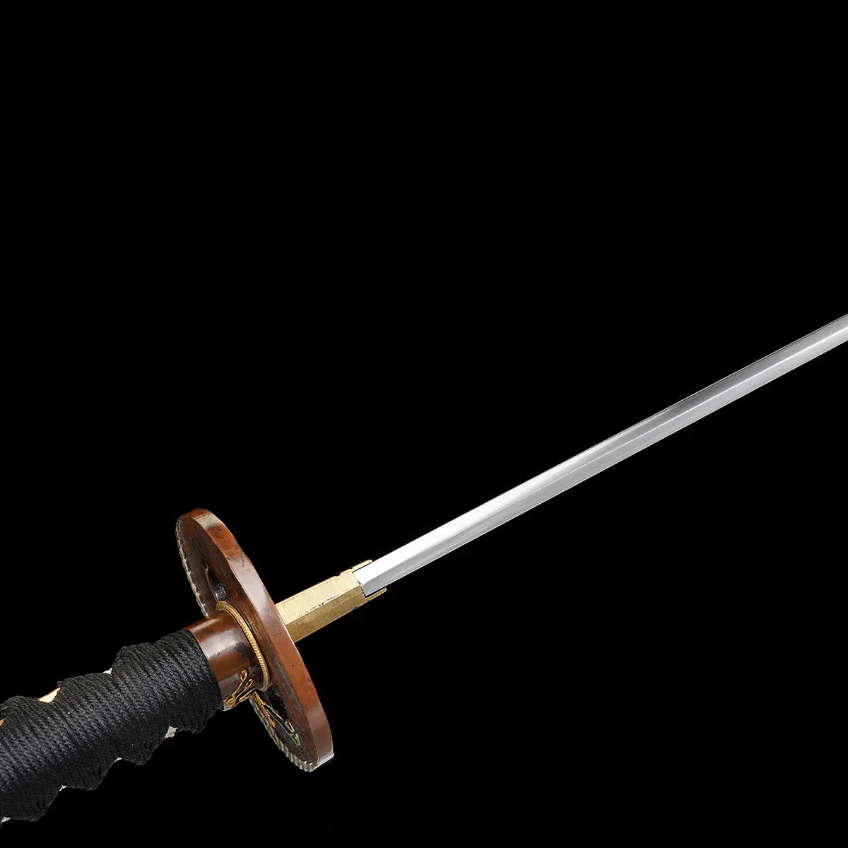 Gros plan sur la poignée du katana japonais artisanal Ryū no Kiba sur fond noir uni, tsuka enveloppée de cordage noir avec losanges blancs en peau de raie, garde tsuba ronde en métal patiné avec motifs décoratifs dorés, habaki doré visible, lame partiellement floutée en arrière-plan sans fourreau ni support.
