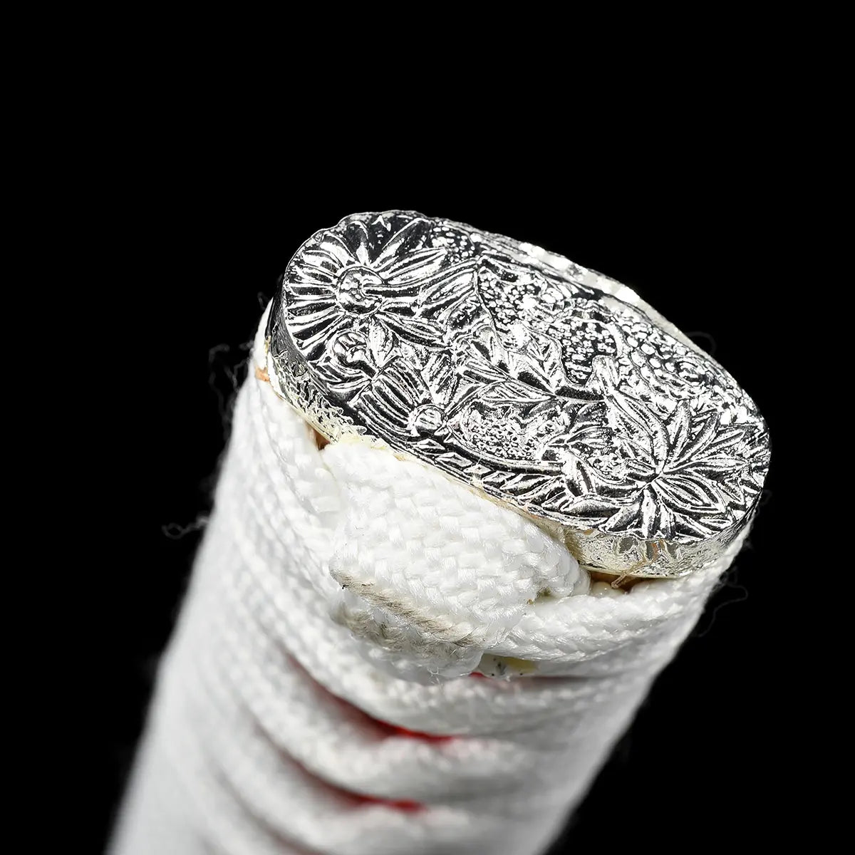 Close-up detail of the end of the handle of the Japanese artisanal katana Hana no Yuki on a solid black background, showing the silver-colored kashira engraved with floral motifs, white tsuka wrapping in white ray skin with white cord knot, subtle red ito visible beneath the wrap, and fine metal detailing at the handle cap

