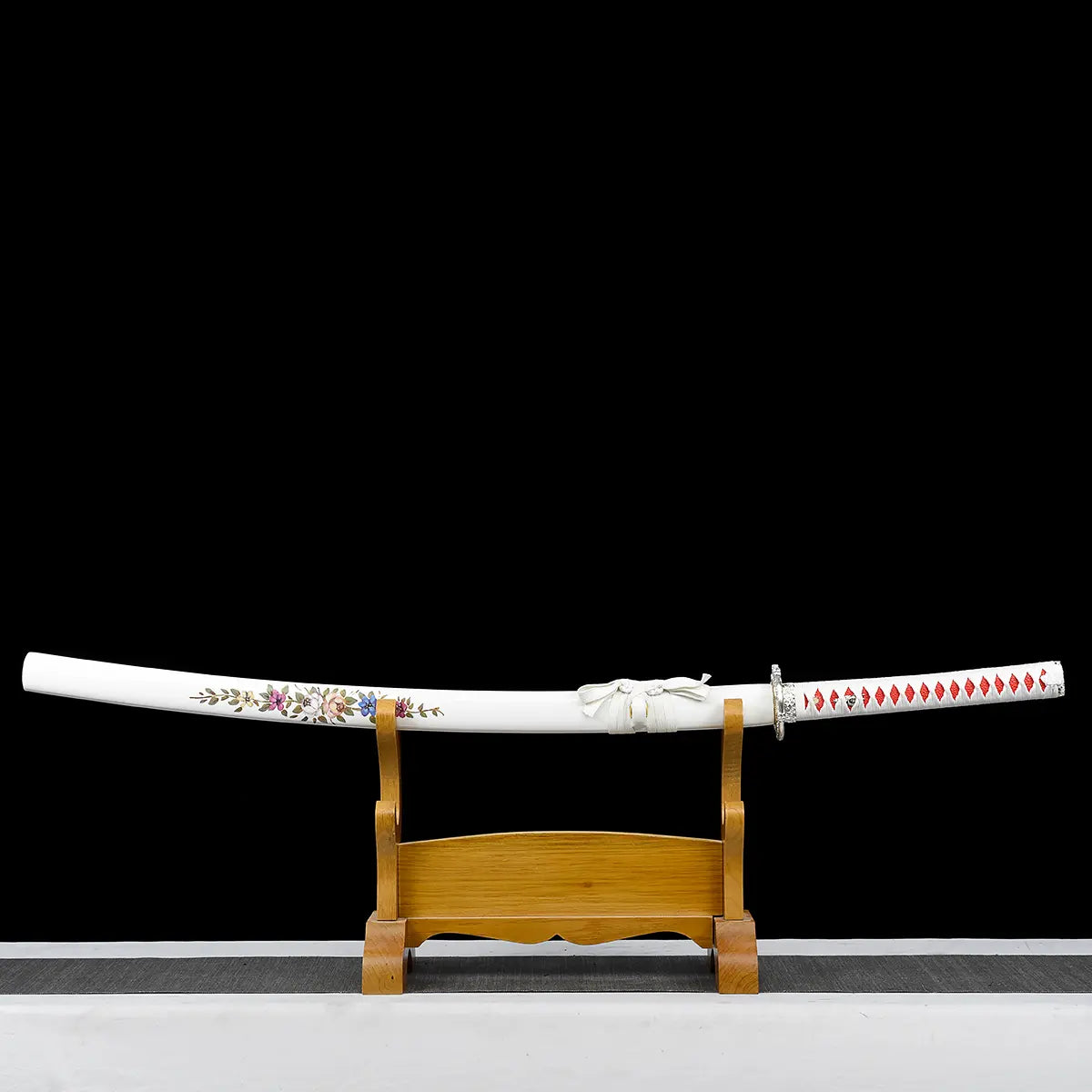 Japanese artisanal katana Hana no Yuki displayed horizontally on a natural wooden stand against a solid black background, featuring a white lacquered saya with delicate multicolored floral patterns, a white tsuka wrapped in white ray skin with red ito diamonds, silver floral tsuba, and the sword fully sheathed resting on the stand
