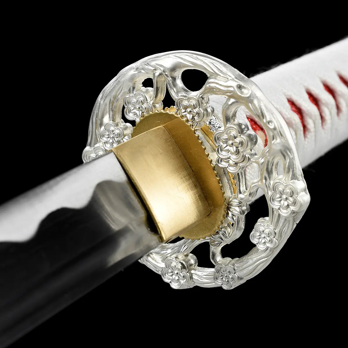 Close-up detail of the tsuba area of the Japanese artisanal katana Hana no Yuki on a solid black background, showing an ornate silver-colored openwork tsuba with floral motifs, a gold-colored brass habaki fitted at the base of the blade, part of the polished steel blade with subtle hamon, and a white tsuka wrapped in white ray skin with red ito diamond pattern
