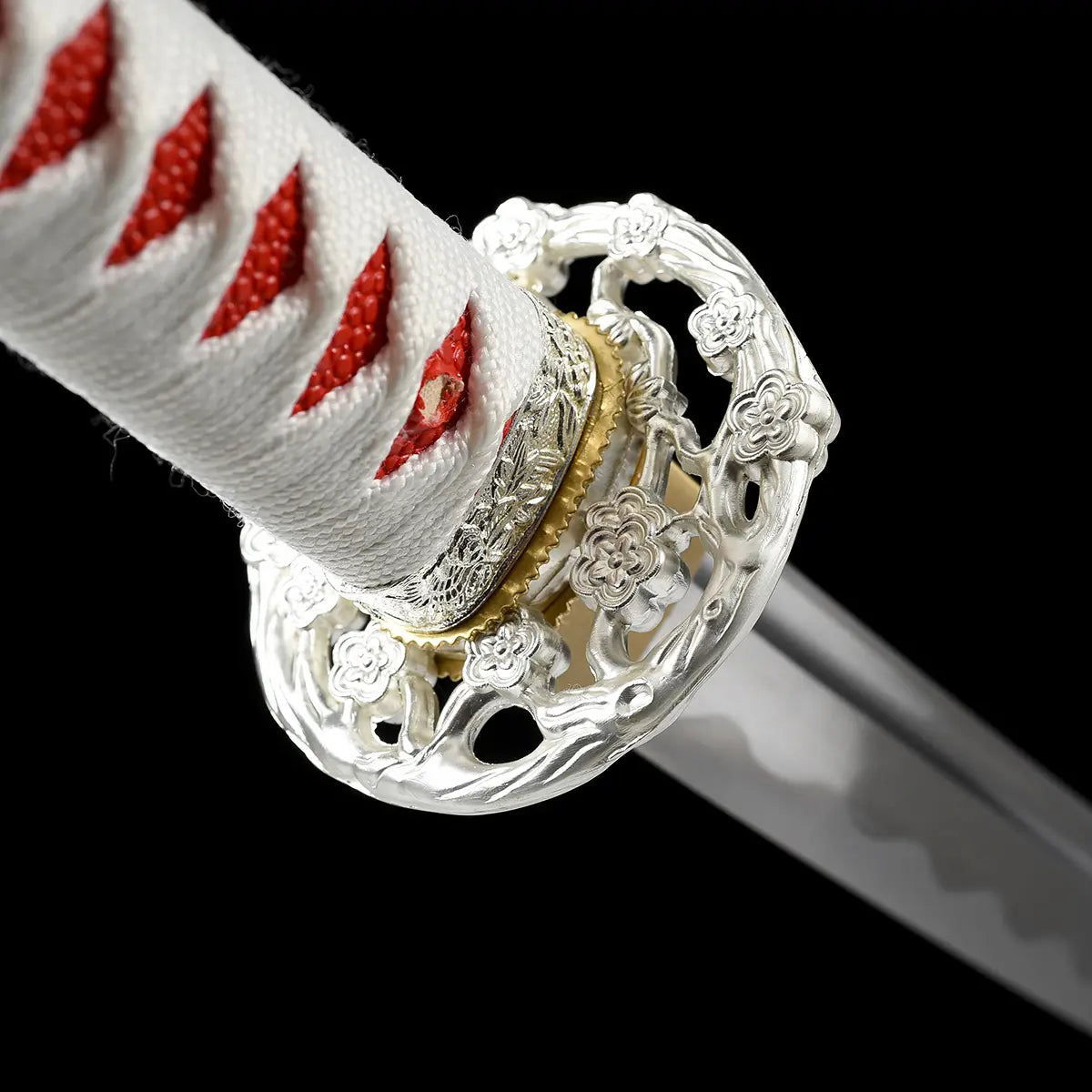 Close-up detail of the handle area of the Japanese artisanal katana Hana no Yuki on a solid black background, showing a white tsuka wrapped in white ray skin with red ito diamond pattern, an ornate silver floral tsuba with openwork design, a gold-colored brass habaki, and the polished steel blade partially visible with a subtle hamon line
