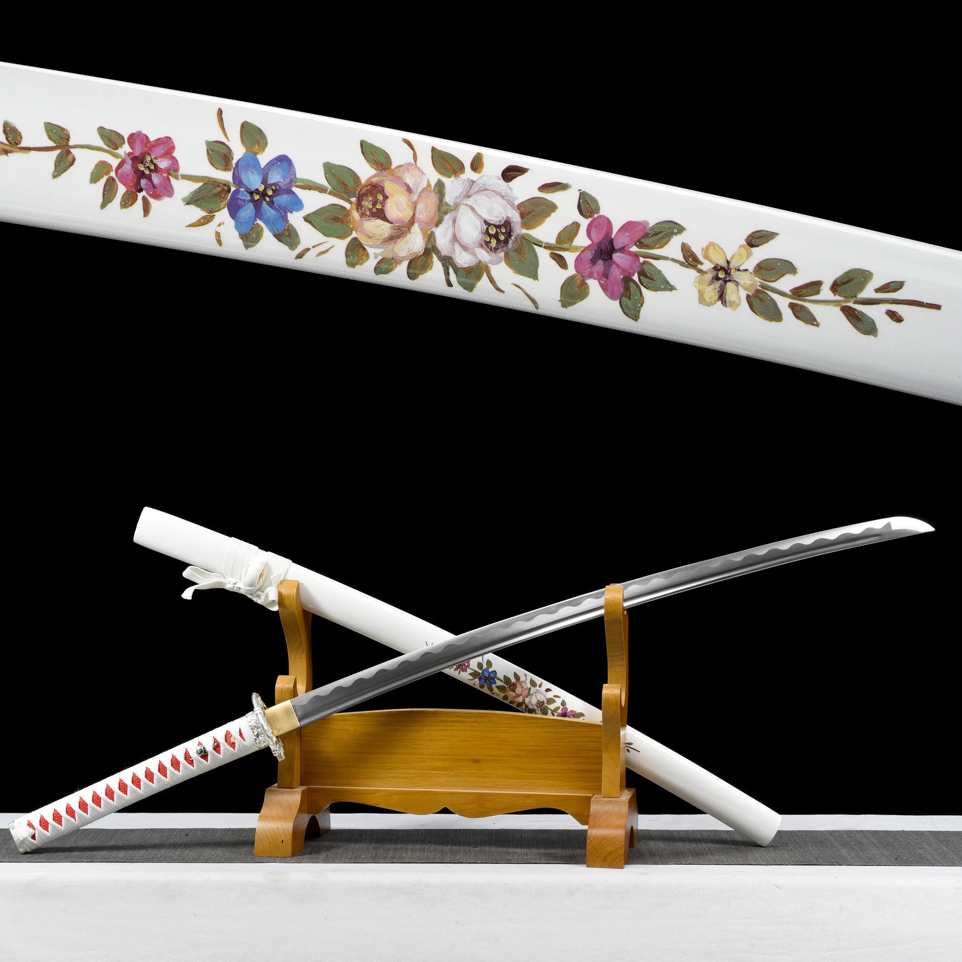 Japanese artisanal katana Hana no Yuki displayed partially unsheathed on a natural wooden stand against a solid black background, showing a polished steel blade with visible bo-hi, a white lacquered saya decorated with detailed multicolored floral motifs, a white tsuka wrapped in white ray skin with red ito diamonds, and a silver floral tsuba, with the blade resting diagonally across the stand
