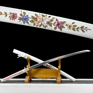 Japanese artisanal katana Hana no Yuki displayed partially unsheathed on a natural wooden stand against a solid black background, showing a polished steel blade with visible bo-hi, a white lacquered saya decorated with detailed multicolored floral motifs, a white tsuka wrapped in white ray skin with red ito diamonds, and a silver floral tsuba, with the blade resting diagonally across the stand

