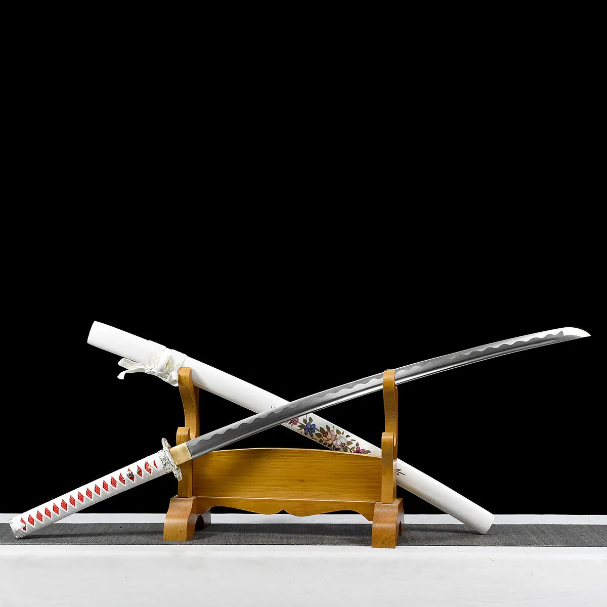 Japanese artisanal katana Hana no Yuki displayed unsheathed in a diagonal position on a natural wooden stand against a solid black background, showing a polished steel blade with visible bo-hi, a white lacquered saya with hand-painted multicolored floral patterns placed behind the blade, a white tsuka wrapped in white ray skin with red ito diamonds, and a silver floral tsuba, with the sword resting securely on the stand
