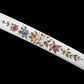 Close-up detail of the white lacquered saya of the Japanese artisanal katana Hana no Yuki on a solid black background, highlighting hand-painted multicolored floral motifs with green leaves, pink, blue, yellow, and white flowers, smooth glossy surface, and the gentle curvature of the scabbard without the blade visible
