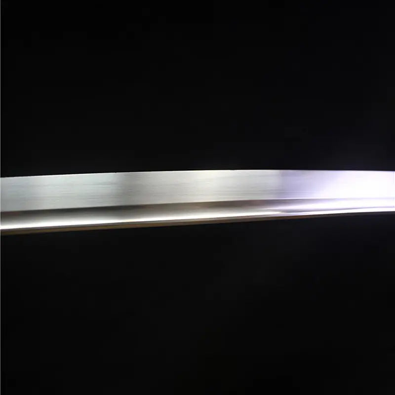 Close-up view of the polished carbon steel blade of the Japanese artisanal katana Mushin, showing smooth linear grain from hand polishing, clean edge line without visible hamon, subtle light reflection along the curvature, deep black background emphasizing the minimalist shirasaya blade finish
