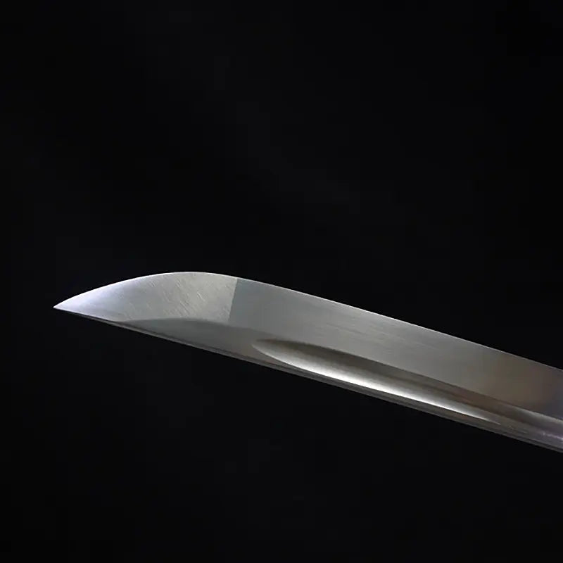 Close-up of the kissaki tip of the Japanese artisanal katana Mushin, showing the polished carbon steel blade with smooth curvature and clean edge line, no hamon visible, satin hand-polished finish, black background highlighting the sharp point and minimalist shirasaya blade profile
