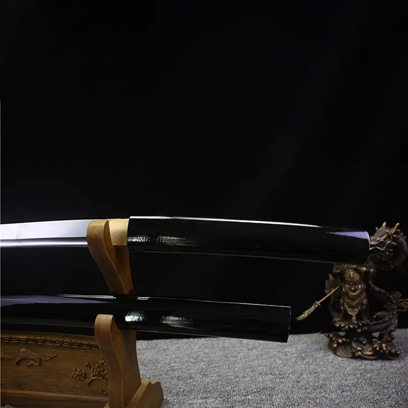 Close-up view of the Japanese artisanal katana Mushin in shirasaya mount, showing the black lacquered wooden saya aligned with the polished steel blade, brass habaki visible near the junction, smooth reflective surface, dark black background and grey fabric base, minimalist and traditional Japanese sword presentation
