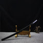 Japanese artisanal katana Mushin displayed in shirasaya mount, black lacquered wooden saya resting diagonally on a wooden sword stand, polished steel blade partially visible, plain brass habaki, dark black background with grey fabric surface, minimalist and traditional presentation
