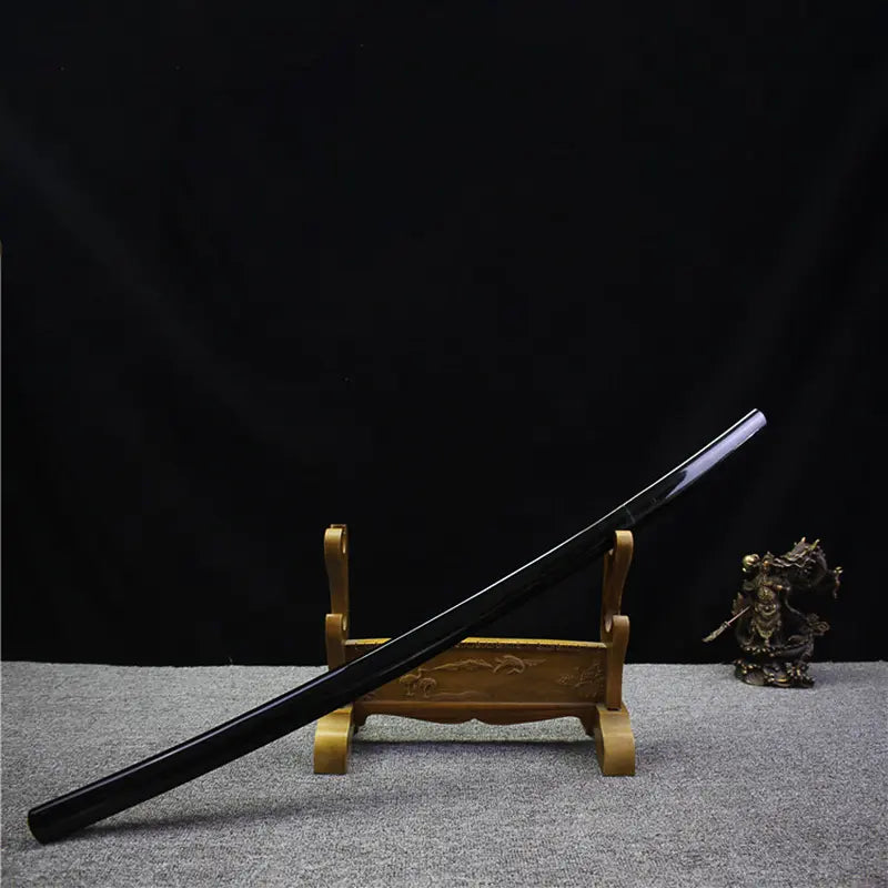 Japanese artisanal katana Mushin displayed in shirasaya mount, black lacquered wooden saya resting diagonally on a wooden sword stand, polished steel blade partially visible, plain brass habaki, dark black background with grey fabric surface, minimalist and traditional presentation
