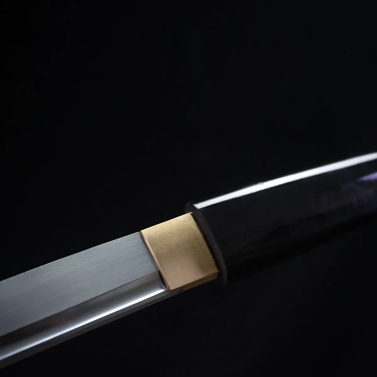 Close-up detail of the Japanese artisanal katana Mushin showing the brass habaki fitted between the polished carbon steel blade and the black lacquered wooden saya, clean junction and smooth surfaces, minimalist shirasaya construction highlighted against a deep black background

