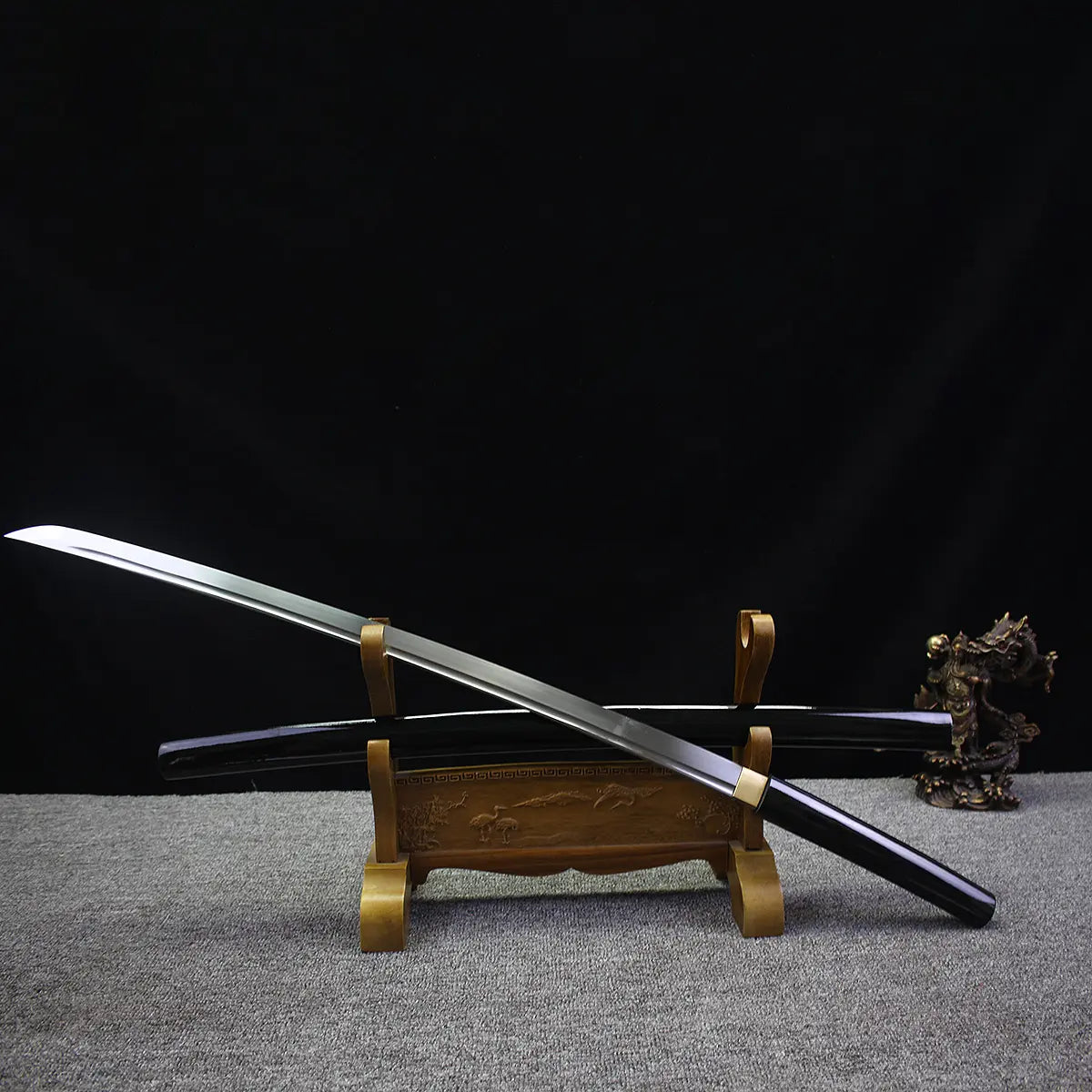 Japanese artisanal katana Mushin displayed diagonally with polished steel blade crossing over its black lacquered wooden saya, both resting on a carved wooden sword stand, brass habaki visible, pointed kissaki highlighted, dark black background with grey fabric surface, minimalist shirasaya composition emphasizing balance and precision
