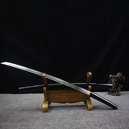 Japanese artisanal katana Mushin displayed diagonally with polished steel blade crossing over its black lacquered wooden saya, both resting on a carved wooden sword stand, brass habaki visible, pointed kissaki highlighted, dark black background with grey fabric surface, minimalist shirasaya composition emphasizing balance and precision

