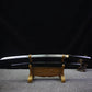 Japanese artisanal katana Mushin displayed horizontally on a carved wooden sword stand, polished steel blade fully visible with smooth curvature, black lacquered wooden saya placed below, brass habaki visible near the handle, dark black background and grey fabric surface, sober and traditional shirasaya presentation
