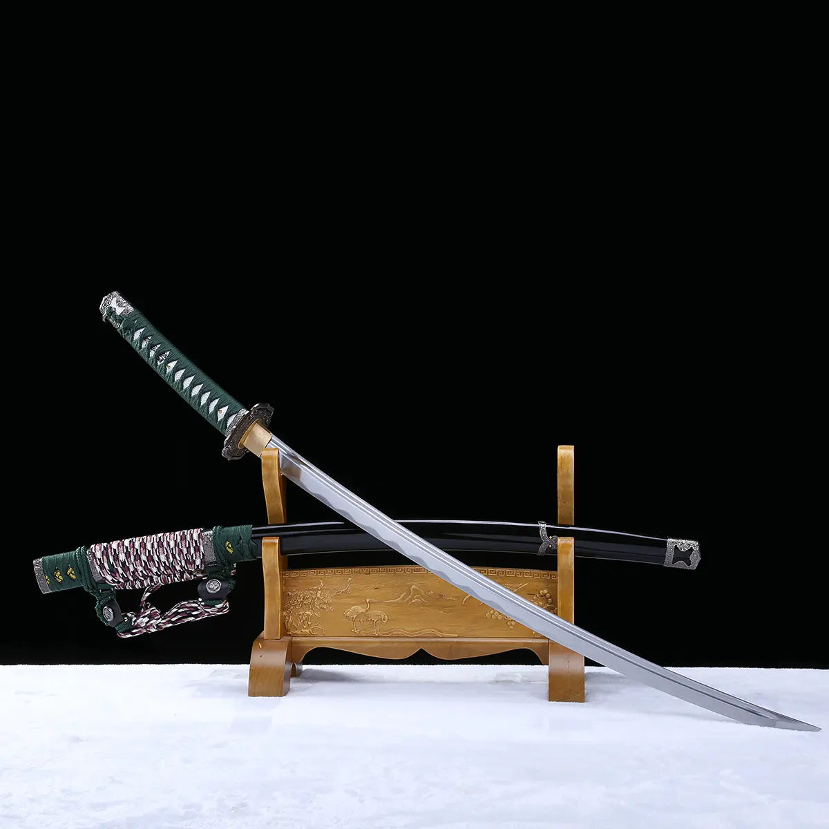 Katana japonais artisanal Seiryū no Tachi photographié sur fond noir avec surface claire, présenté en diagonale sur un support en bois sculpté. La lame courbe en acier carbone est sortie du fourreau et orientée vers le bas, laissant apparaître un hamon réel ondulé sur toute la longueur. Le fourreau noir laqué repose à l’arrière sur le support, avec son sageo tressé vert, blanc et pourpre. Le manche est recouvert d’un tsuka-ito vert foncé avec same blanche apparente en losanges, et les montures métalliques t