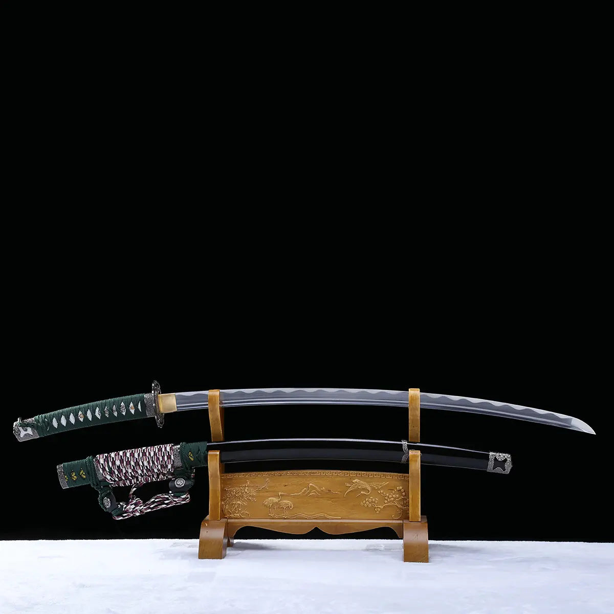 Katana japonais artisanal Seiryū no Tachi photographié sur fond noir avec base claire, présenté en pleine longueur sur un support en bois sculpté. La lame courbe en acier carbone est entièrement visible hors du fourreau, révélant un hamon réel ondulé sur toute la longueur. Le fourreau noir laqué est positionné sous la lame, avec son sageo tressé vert, blanc et pourpre. Le manche est recouvert d’un tsuka-ito vert foncé laissant apparaître une same blanche en losanges, et les montures métalliques traditionnel