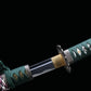 Détail de la base de la lame du katana japonais artisanal Seiryū no Tachi photographié sur fond noir uni. La lame en acier carbone est partiellement sortie du fourreau, montrant un habaki de couleur laiton ajusté avec précision. La transition entre la lame polie et le manche est visible, avec une garde métallique sculptée, un tsuka-ito vert foncé tressé serré et une same blanche en losanges apparente s
