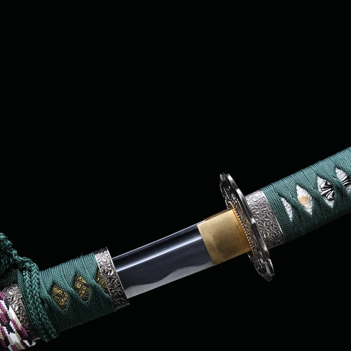 Détail de la base de la lame du katana japonais artisanal Seiryū no Tachi photographié sur fond noir uni. La lame en acier carbone est partiellement sortie du fourreau, montrant un habaki de couleur laiton ajusté avec précision. La transition entre la lame polie et le manche est visible, avec une garde métallique sculptée, un tsuka-ito vert foncé tressé serré et une same blanche en losanges apparente s
