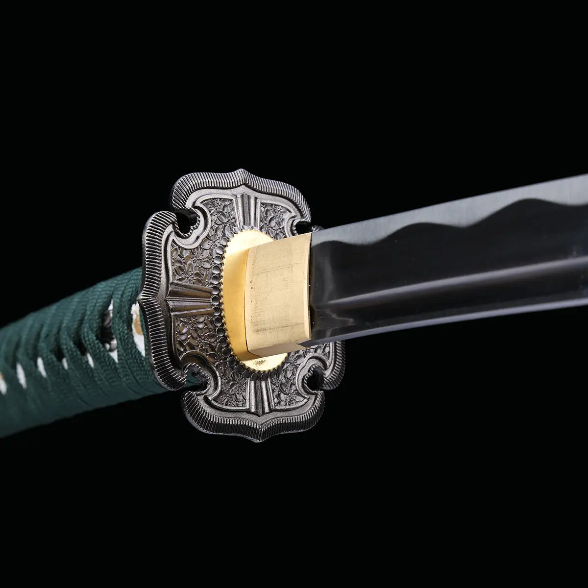 Détail rapproché de la tsuba et de la base de la lame du katana japonais artisanal Seiryū no Tachi photographié sur fond noir uni. La garde métallique finement sculptée présente des motifs floraux en relief, associée à un habaki de couleur laiton parfaitement ajusté. La lame courbe en acier carbone est visible avec un polissage net et un début de hamon ondulé, tandis que le manche recouvert d’un tsuka-ito vert foncé apparaît partiellement en arrière-plan.
