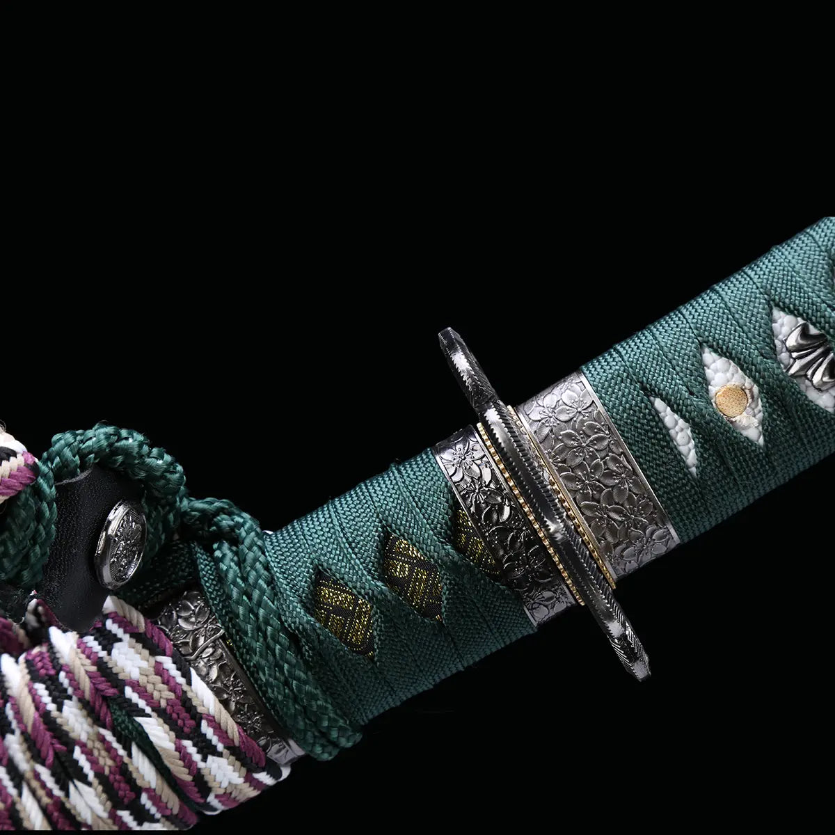 Détail de la zone tsuba et habaki du katana japonais artisanal Seiryū no Tachi photographié sur fond noir uni. La garde métallique sculptée de motifs floraux est visible, associée à un habaki de teinte laiton. Le manche est recouvert d’un tsuka-ito vert foncé tressé, laissant apparaître une same blanche en losanges, avec menuki métalliques décoratifs intégrés sous le tressage. Le sageo multicolore est attaché à la base du manche.
